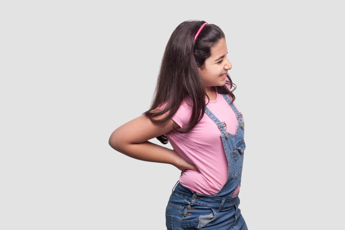 Spine back pain. Profile side view Portrait of sick brunette young girl in pink t-shirt and blue overalls standing and holding her painful back.
