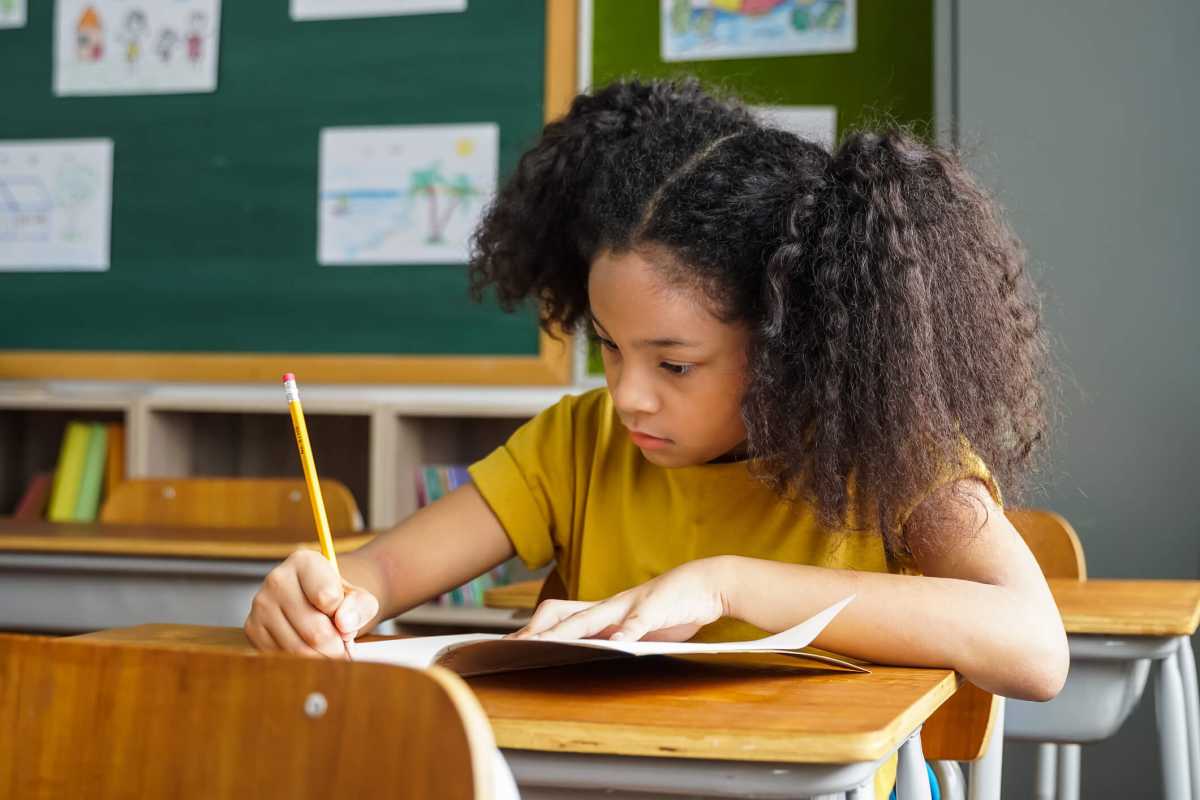 African American school girl sitting in school writing in book with pencil, studying, education, learning. Female student sitting at desk in classroom writing in notebook in exam