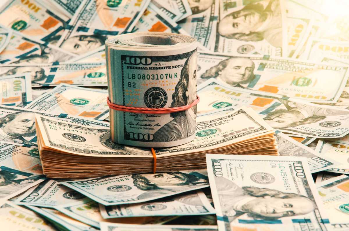 A background of scattered one-hundred-dollar bills over the entire space, close-up in the middle of a stack and a roll of dollars in the shape of a ship.