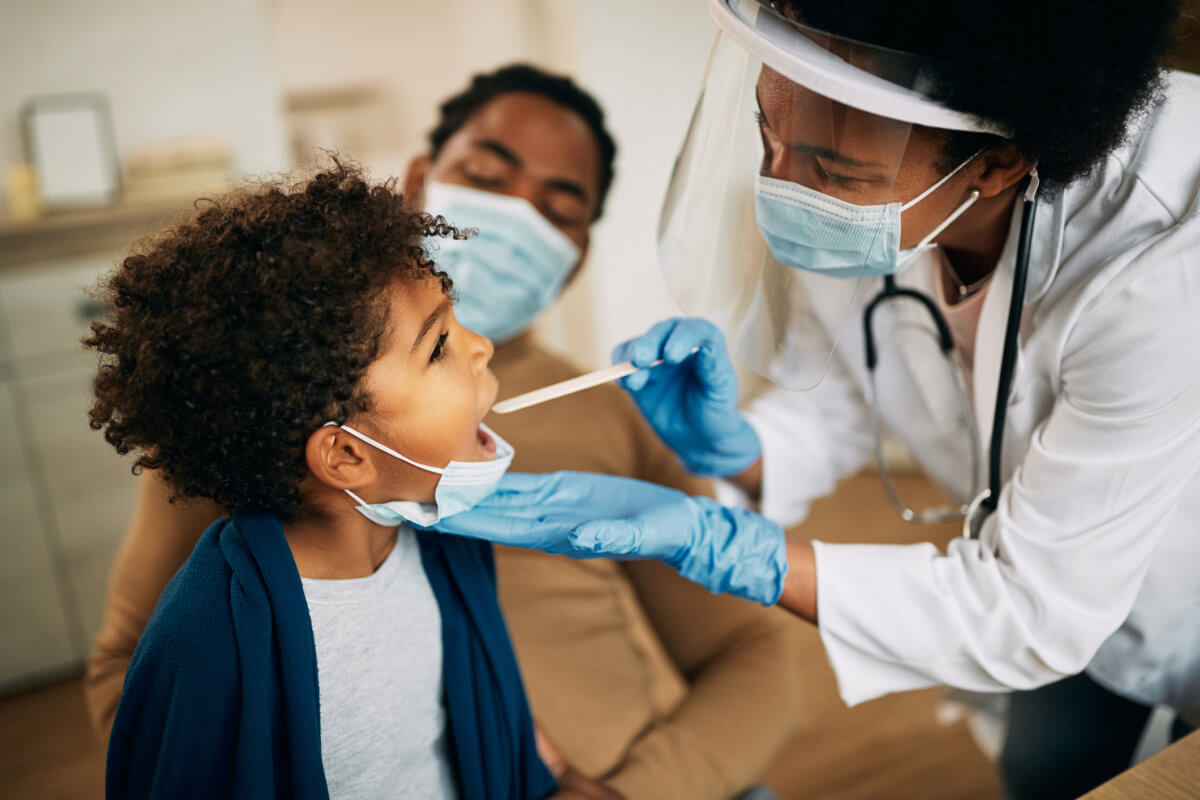 African American doctor with face mask examining boy’s throat during a home visit.
