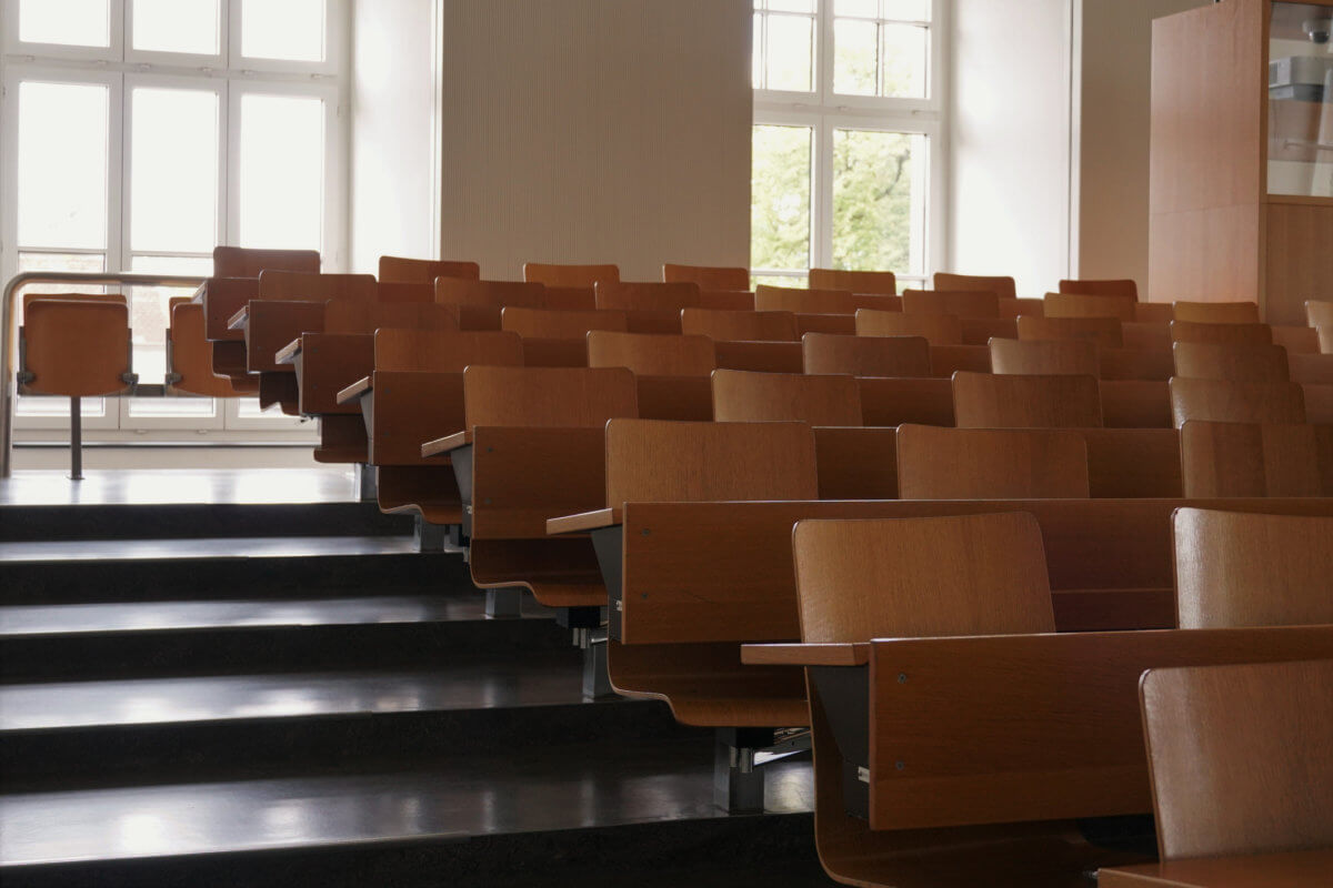 Empty auditorium at university with wooden chairs and banks and large windows and stairs on one side.