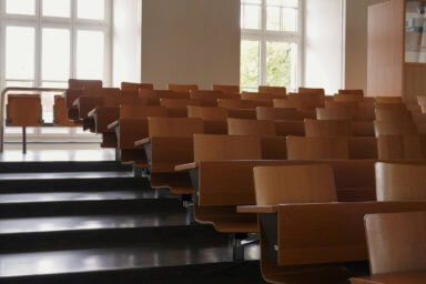 Empty auditorium at university with wooden chairs and banks and large windows and stairs on one side.