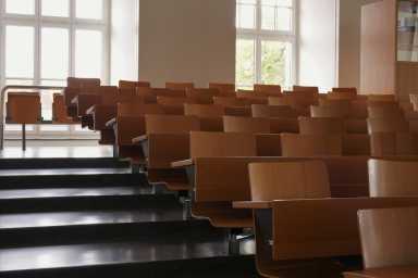 Empty auditorium at university with wooden chairs and banks and large windows and stairs on one side.