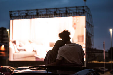 Silhouetted view of attractive young couple, boy and girl embracing, spending time together, sitting in the car while watching a movie in a drive in cinema