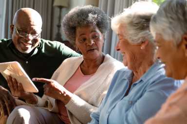 Three happy diverse senior woman and african american male friend sitting on sofa and using tablet