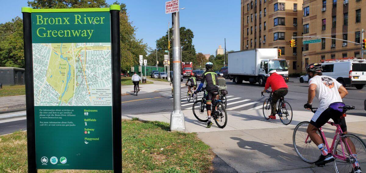 people ride their bicycles by a Bronx River Greenway sign
