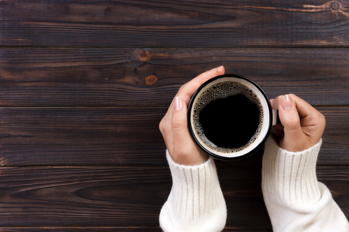 Lonely woman drinking coffee in the morning, top view of female hands holding cup of hot beverage on wooden desk