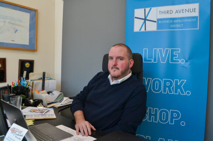 Michael Brady sitting in an office chair in front of a Third Avenue BID sign that says Live. Work. Shop.