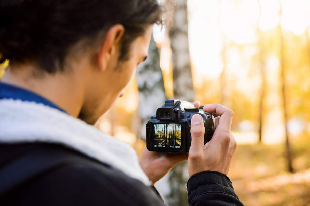 Back view of a photograper shooting pictures on a DSLR camera, blurred background of trees.