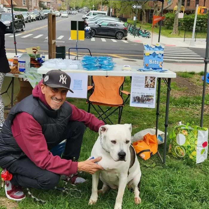 Guy crouching down beside his white pitbull on some grass in front of a table.