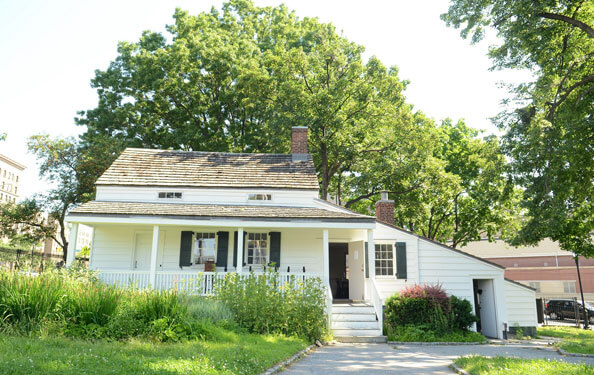 Edgar Allan Poe's cottage surrounded by trees, grass and bushes on a sunny day.