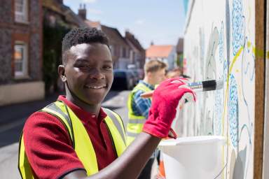 Teenagers wearing safety vests paint a mural outdoors on a brick wall
