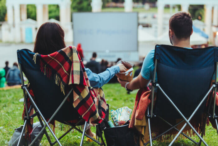 Two people sitting in camp chairs sharing a snack at an outdoor movie showing in a park in daylight. What's Happening.