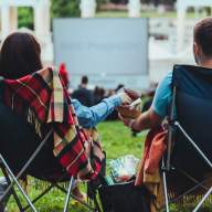Two people sitting in camp chairs sharing a snack at an outdoor movie showing in a park in daylight. What's Happening.