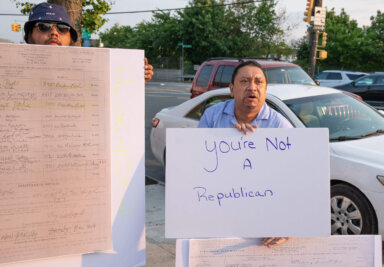 two protestors hold signs. one sign is a picture of a petition and the other sign says "You're not a Republican"