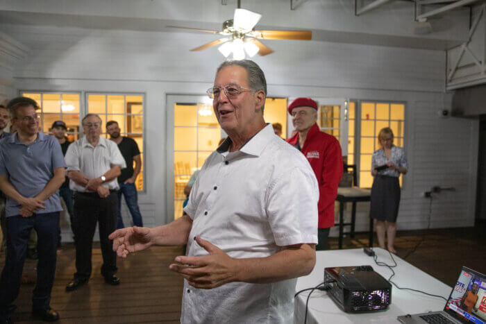 George Havranek speaks. A crowd of supporters as well as George and Nancy Sliwa are behind him. A laptop and projector can be seen on the table.