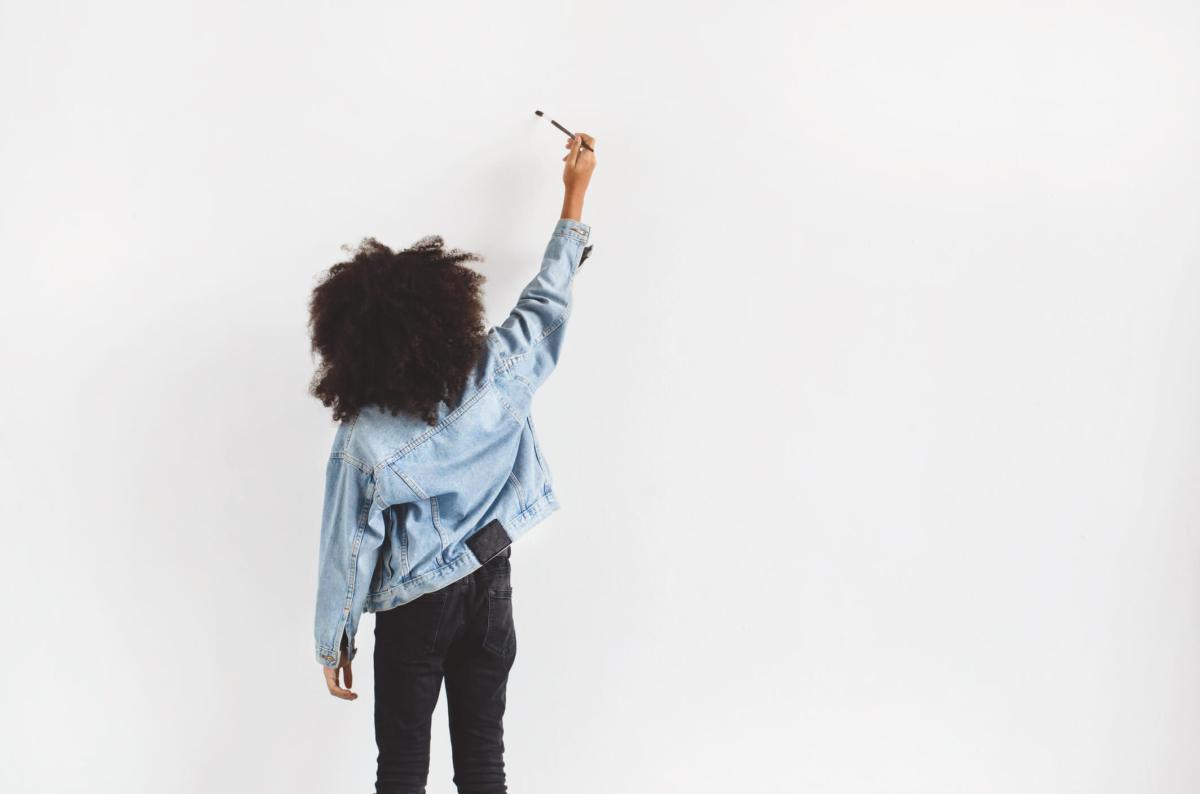 A dark-skinned girl stands near the wall and writes something.