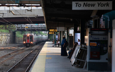 A train approaches on the tracks as passengers wait on the platform. Photo also shows an MTA ticket machine and a sign that says "To New York" on the platform.