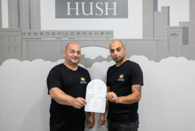 Levent and Denis Ozkurt pose with a sticker to put in their window signalling they are licensed. They stand in front of a mural with a Bronx subway and a sign that says Hush.