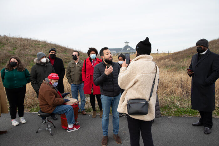 Jonathan Soto speaks while a woman records him on Instagram live. Various people stand behind him, with one person playing the drums. The Trump Golf Links clubhouse is also in the background.