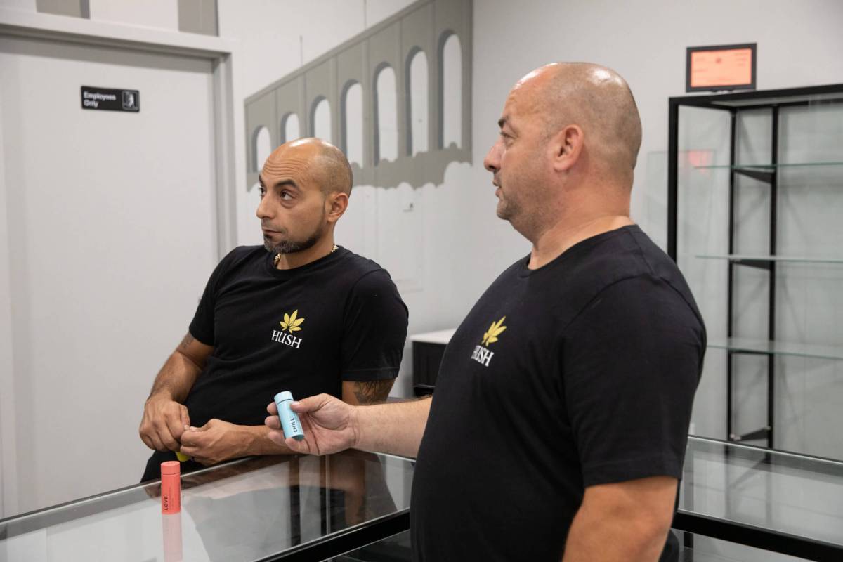 Denis Ozkurt looks off camera with a bottle of cannabis capsules labeled "love" on a counter in front of him and his cousin Levent Ozkurt stands on the other side of the counter holding a bottle that says "chill" on it.