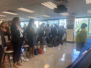 group of new hires standing with their hand up in front of folding chairs with Darcel Clark speaking into a microphone
