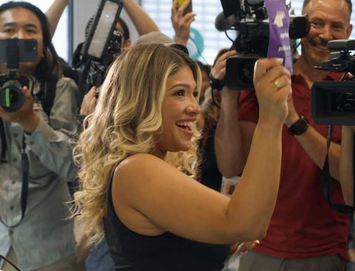 woman smiles with arm up holding a product as people have cameras on her