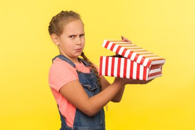 Portrait of upset child opening gift box, little girl in denim overalls looking at camera with disappointed face