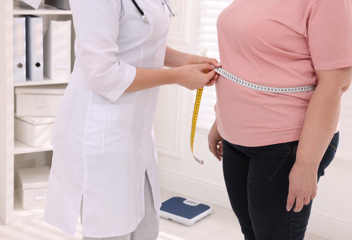 Nutritionist measuring overweight woman’s waist with tape in clinic, closeup