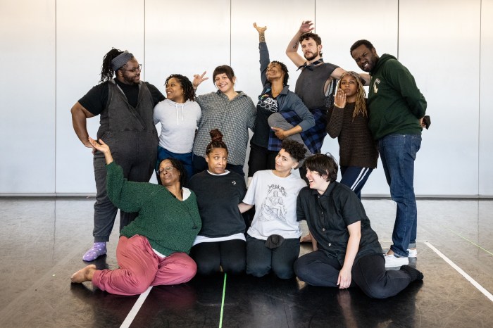 The cast of How to Bend Down/ How to Pick it Up poses for a group photo. Some performers stand and some sit or kneel on the studio floor. It is a joyful moment: everyone is smiling, gesturing, or making a fun face.