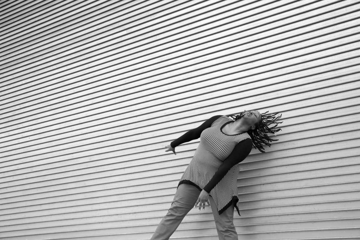 A black & white image of Kayla Hamilton, a dark-skinned Black woman, dancing in front of a textured wall that has horizontal layers. Her arms are energetically reaching down to the diagonal as her head is tilting to the diagonal with her dreads moving back in that same direction. She wears a diagonally striped long sleeve shirt with pants.