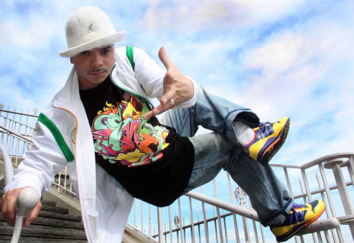 Kwikstep balancing on a staircase railing with one hand and one foot. The hand not on the railing is reaching towards the camera, palm open and fingers extended. He is wearing a white, furry Bell Kango hat, a white and green Adidas jacket, a black t-shirt with colorful graffiti letters, blue jeans, and colorful Adidas sneakers. Photo by B Fresh.